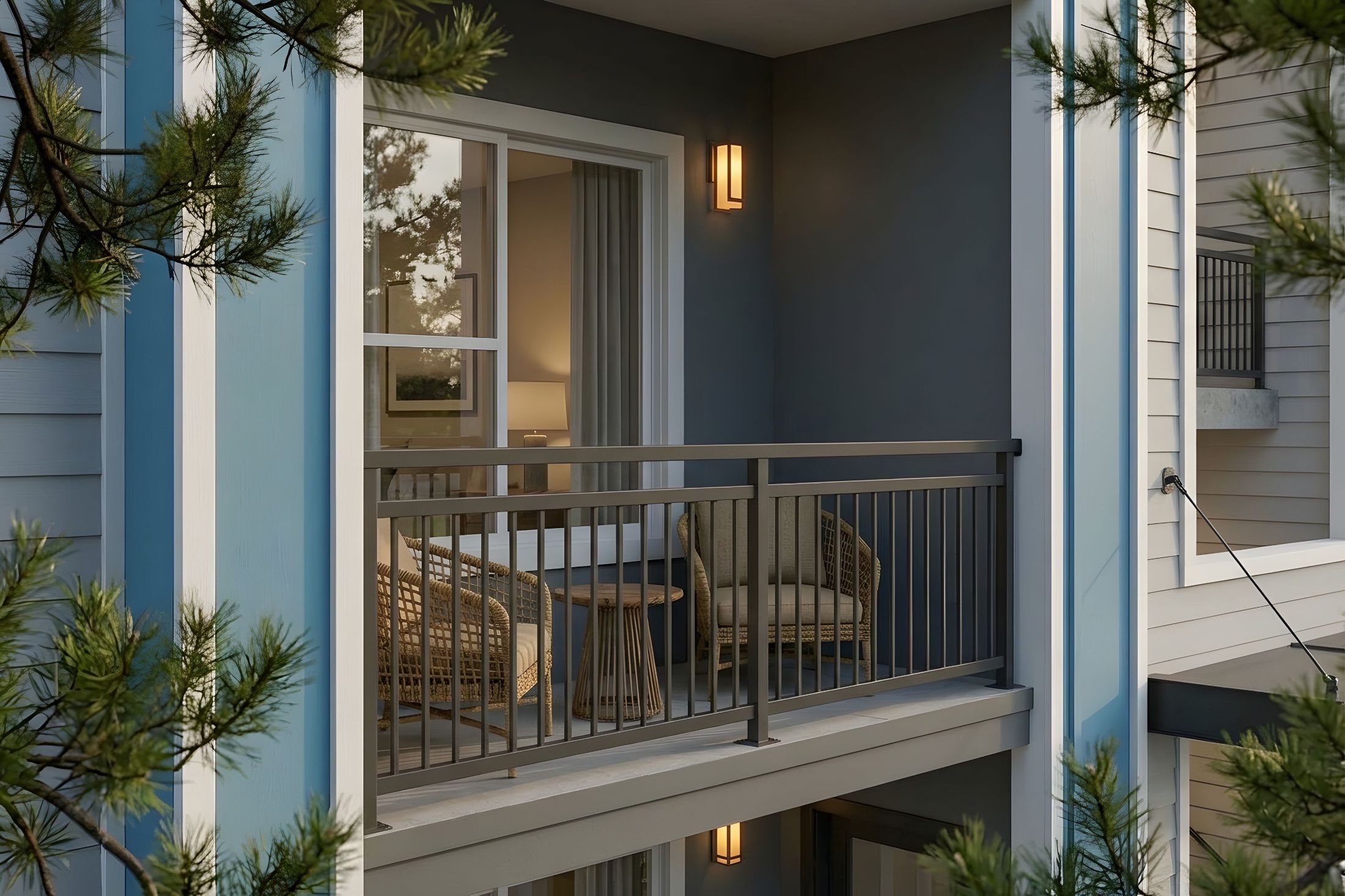 A modern apartment balcony with two wicker chairs and a small wooden table, enclosed by a dark metal railing, illuminated by a warm wall sconce light.